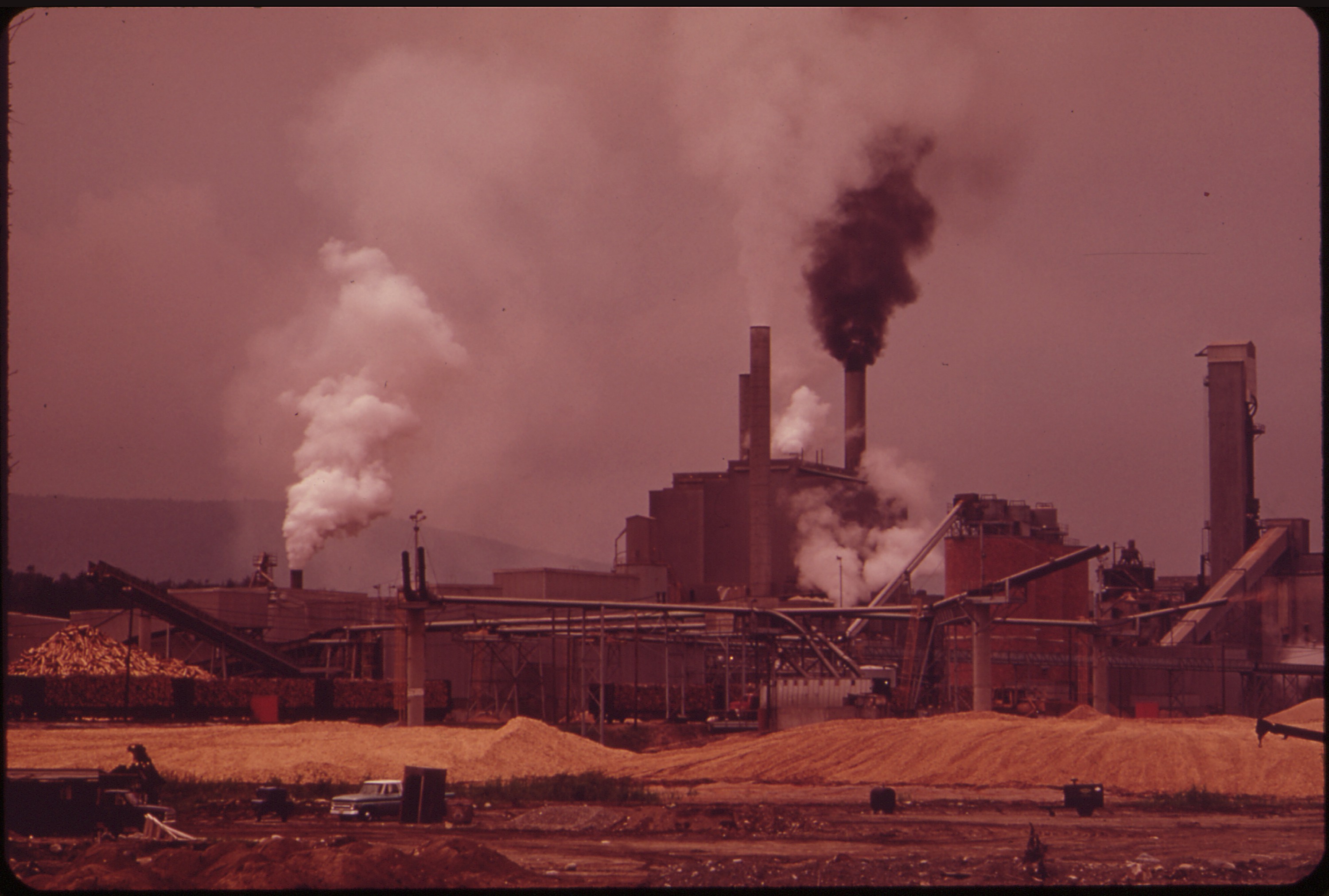 Black-and-orange photograph of the Androscoggin paper mill in Jay, Maine, with smokestacks emitting heavy plumes against a hazy sky.